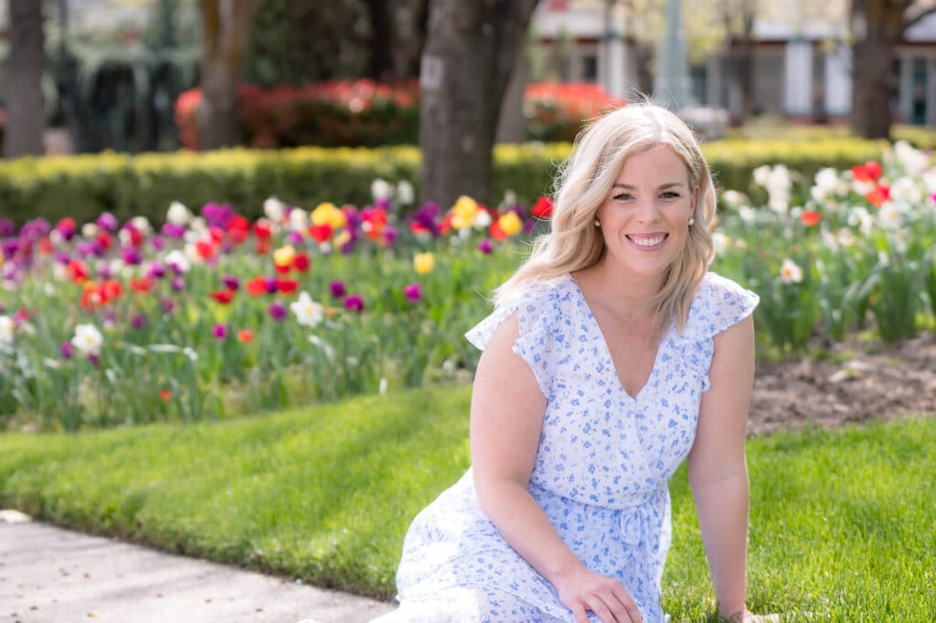 Katie Rouse, founder and owner of Style by Design Co, wearing a white dress, standing in front of a bed of flowers in Boise, Idaho